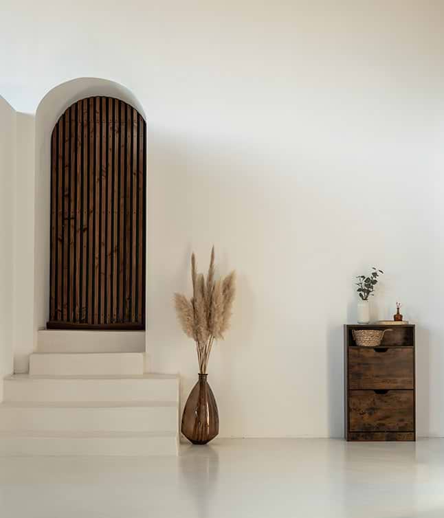 Minimalist interior featuring a wooden arched door, white staircase, pampas grass in a vase, and a rustic drawer cabinet.
