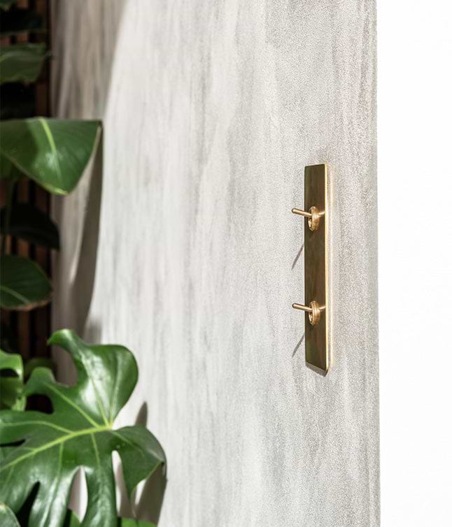 Close-up of a textured gray wall featuring a sleek brass light switch, with a glimpse of a white cabinet and lush greenery.