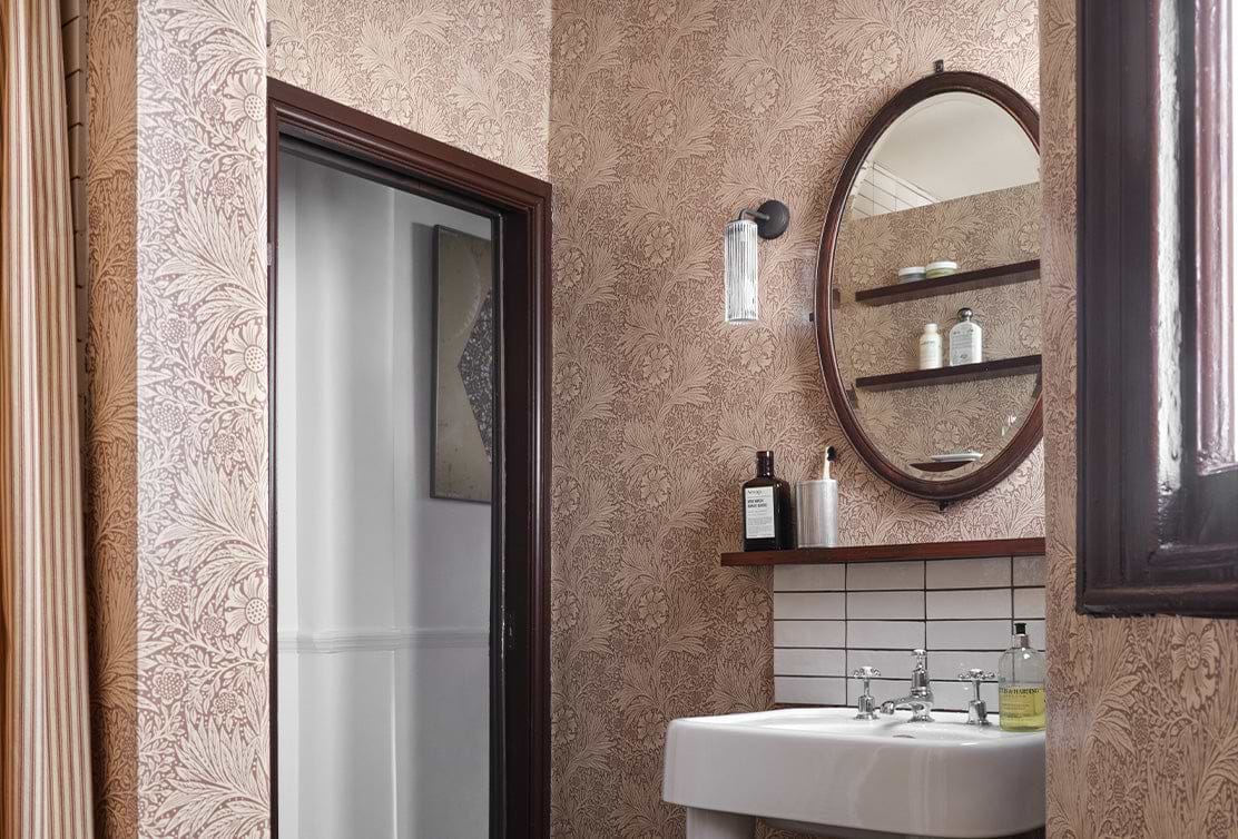 A stylish bathroom featuring floral wallpaper, a round mirror, a glass light fixture, and a minimalist sink with shelving.