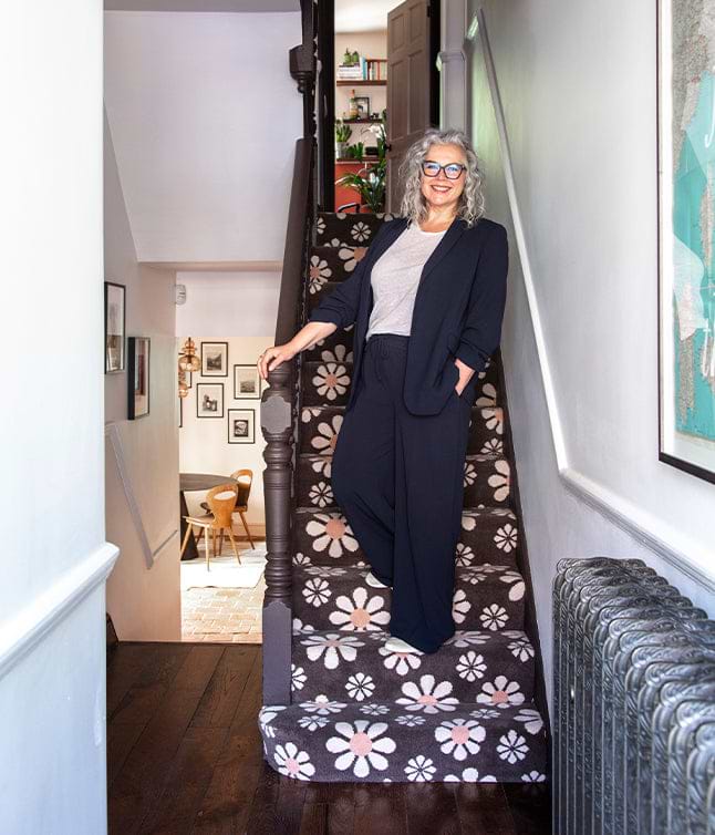 A person in a navy suit and grey top stands on a flower-patterned staircase, leading to a stylish, light-filled room.