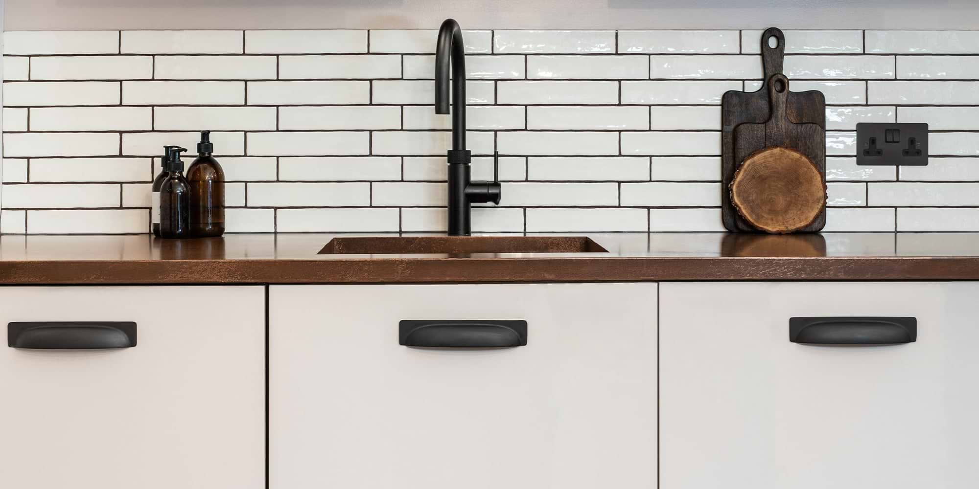 Modern kitchen countertop with a black faucet, glass bottles, wooden cutting board, and white subway tile backsplash.