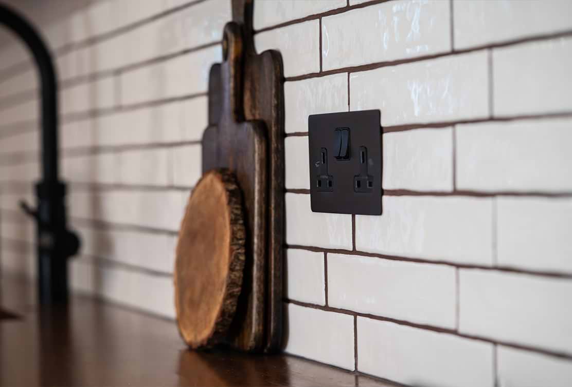 A dark electrical outlet on a white tiled kitchen wall, beside a wooden cutting board with a natural edge.