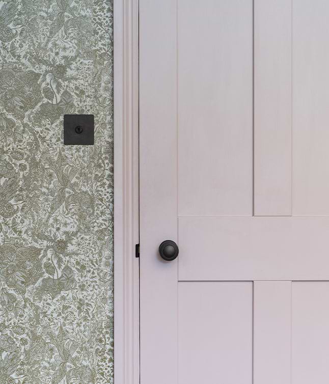 A close-up of a pale pink door with a black knob, adjacent to textured green floral wallpaper.