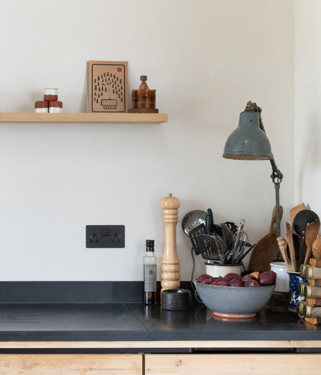 A rustic kitchen counter featuring a bowl of plums, cooking utensils, a pepper grinder, and an industrial lamp against a simple backdrop.