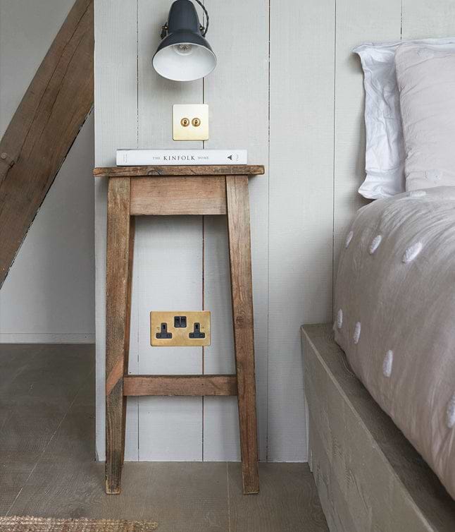 A rustic wooden bedside table holds a book, with a stylish wall lamp above and a gold electrical outlet below in a cozy bedroom setting.