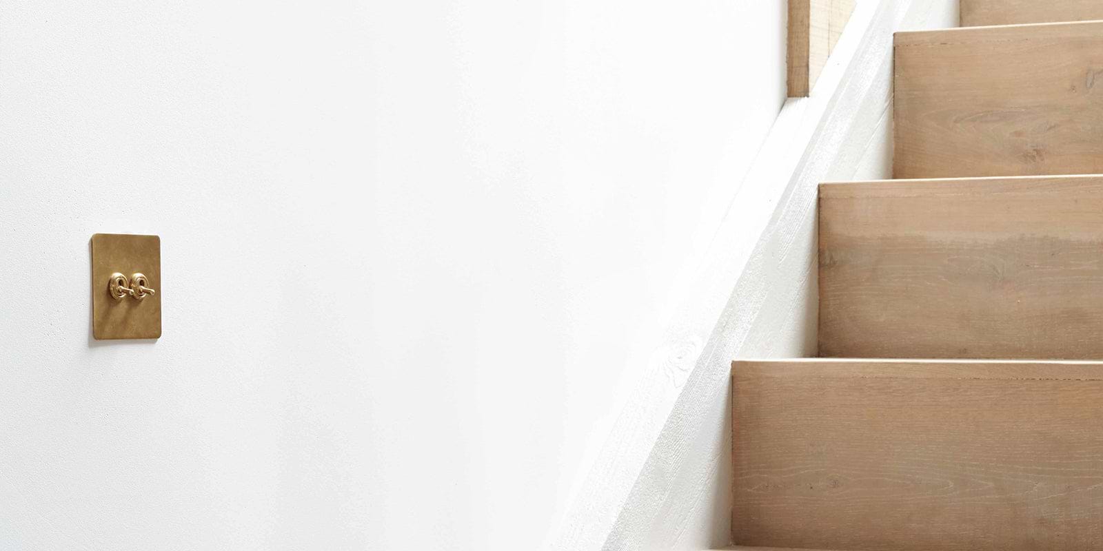 A brass light switch plate mounted on a white wall beside a staircase with light wood steps, creating a modern and minimalistic aesthetic.