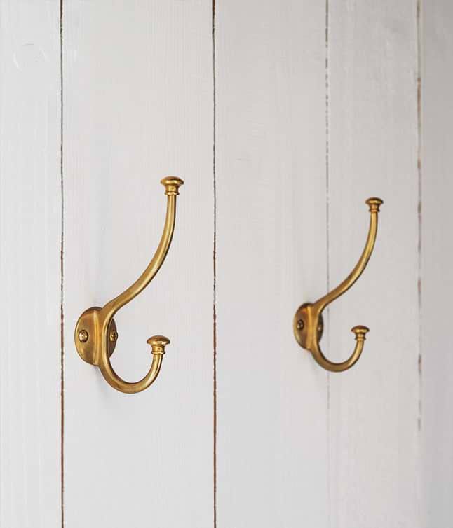 Two elegant brass hooks mounted on a white wooden wall, showcasing a vintage design for hanging items.