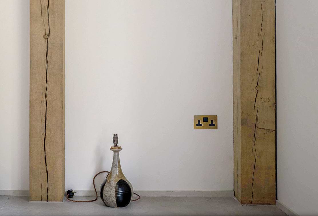 A rustic corner featuring a textured wooden beam, a minimalist ceramic lamp, and a brass electrical outlet against a plain wall.