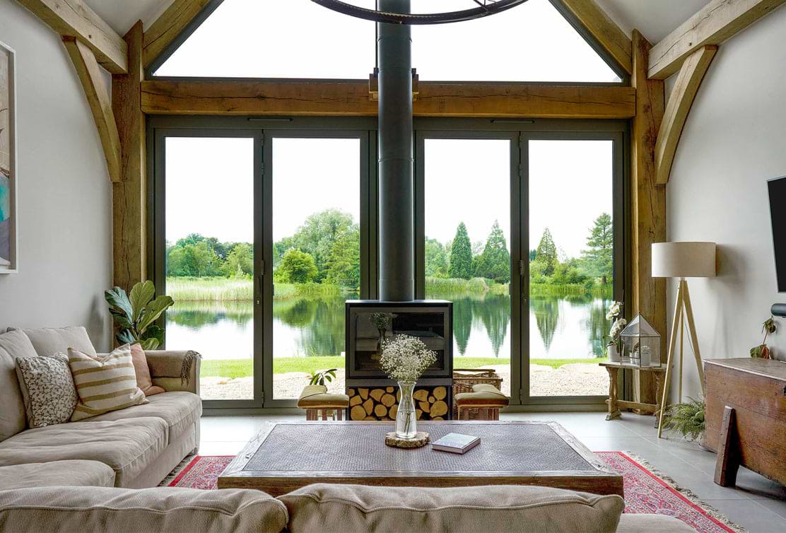 A cozy lakeside living room with a beige sofa, wooden accents, and large windows offering a serene view of the lush green landscape.