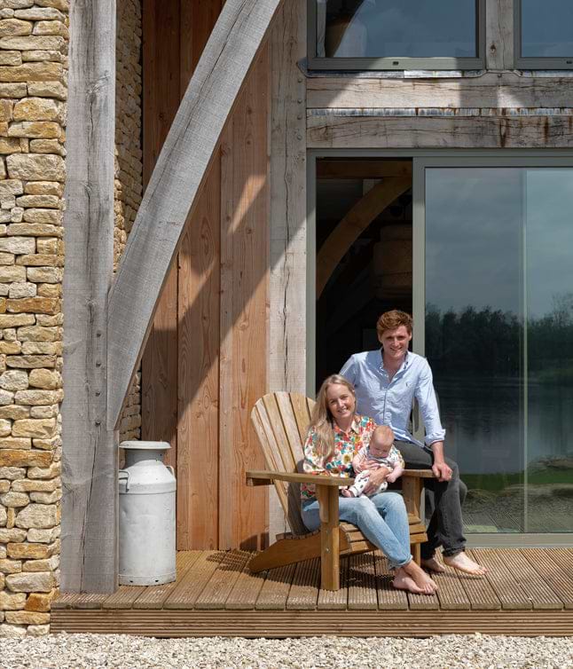 A family enjoys a sunny day on a wooden deck, posing with a baby, with a scenic lakeside view and rustic architecture in the background.