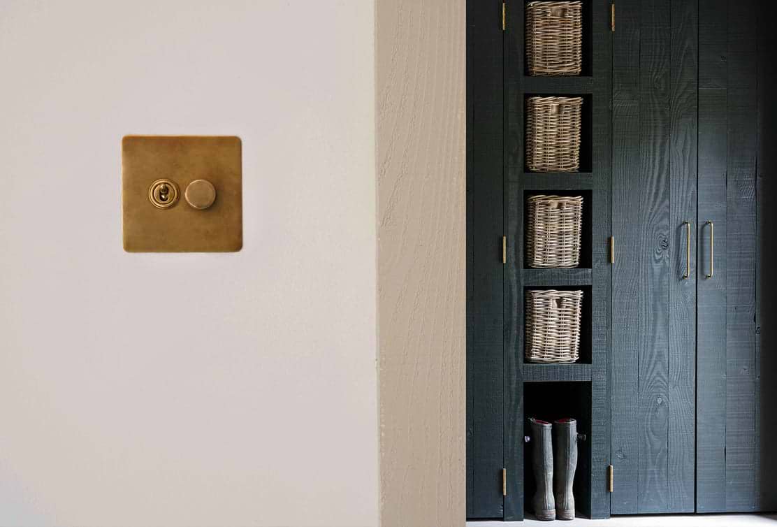 A brass light switch and keyhole on a white wall, beside a dark cabinet with four woven baskets and a pair of gray boots at the bottom.