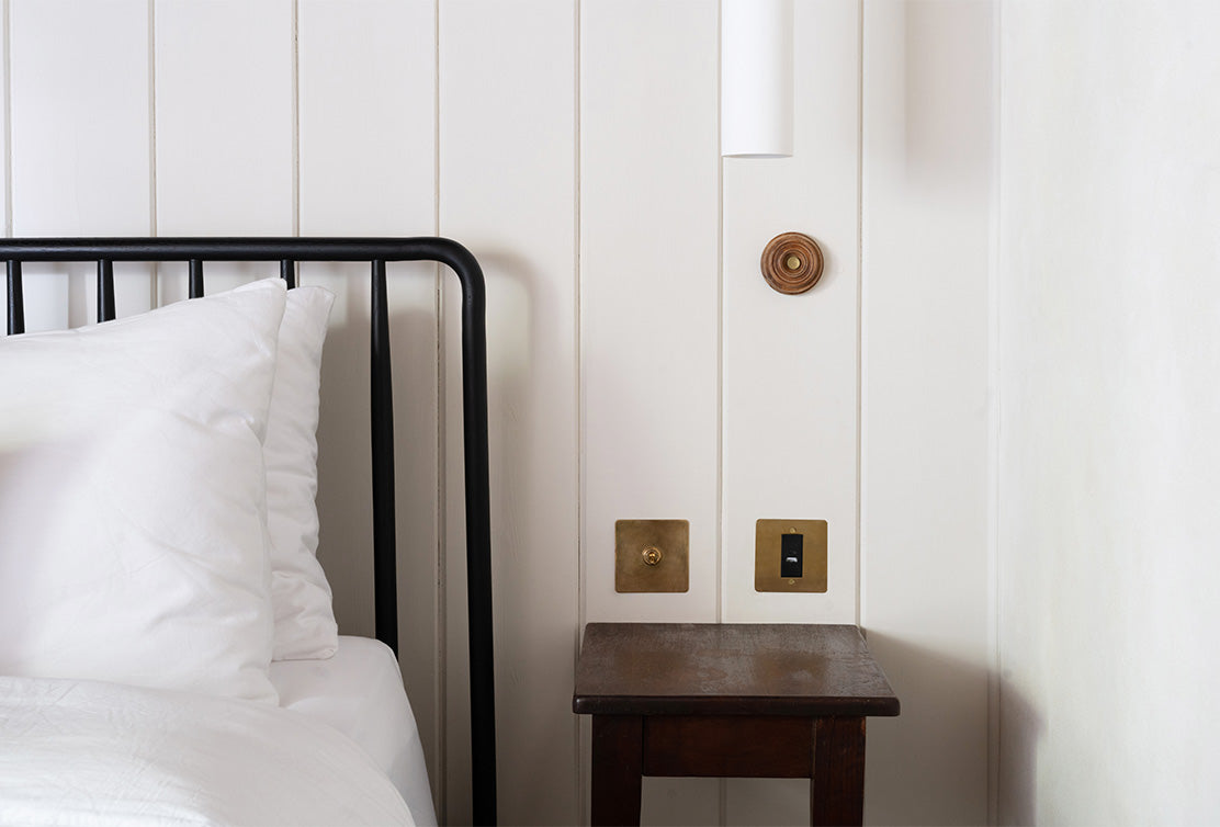 A minimalist bedroom corner featuring a black metal bed frame, white pillows, a small wooden bedside table, and brass wall switches.