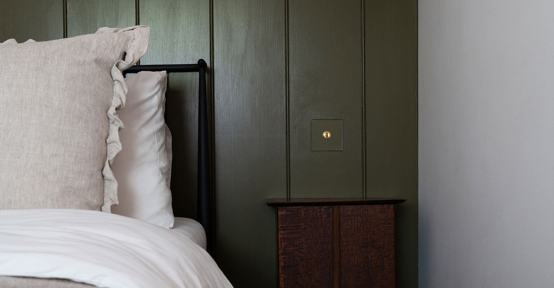 Cozy bedroom corner with a ruffled beige pillow, a sleek black bed frame, and a dark green wall featuring a simple brass light switch.