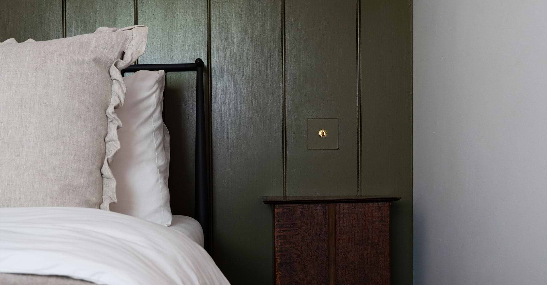 Cozy bedroom corner with a ruffled beige pillow, a sleek black bed frame, and a dark green wall featuring a simple brass light switch.