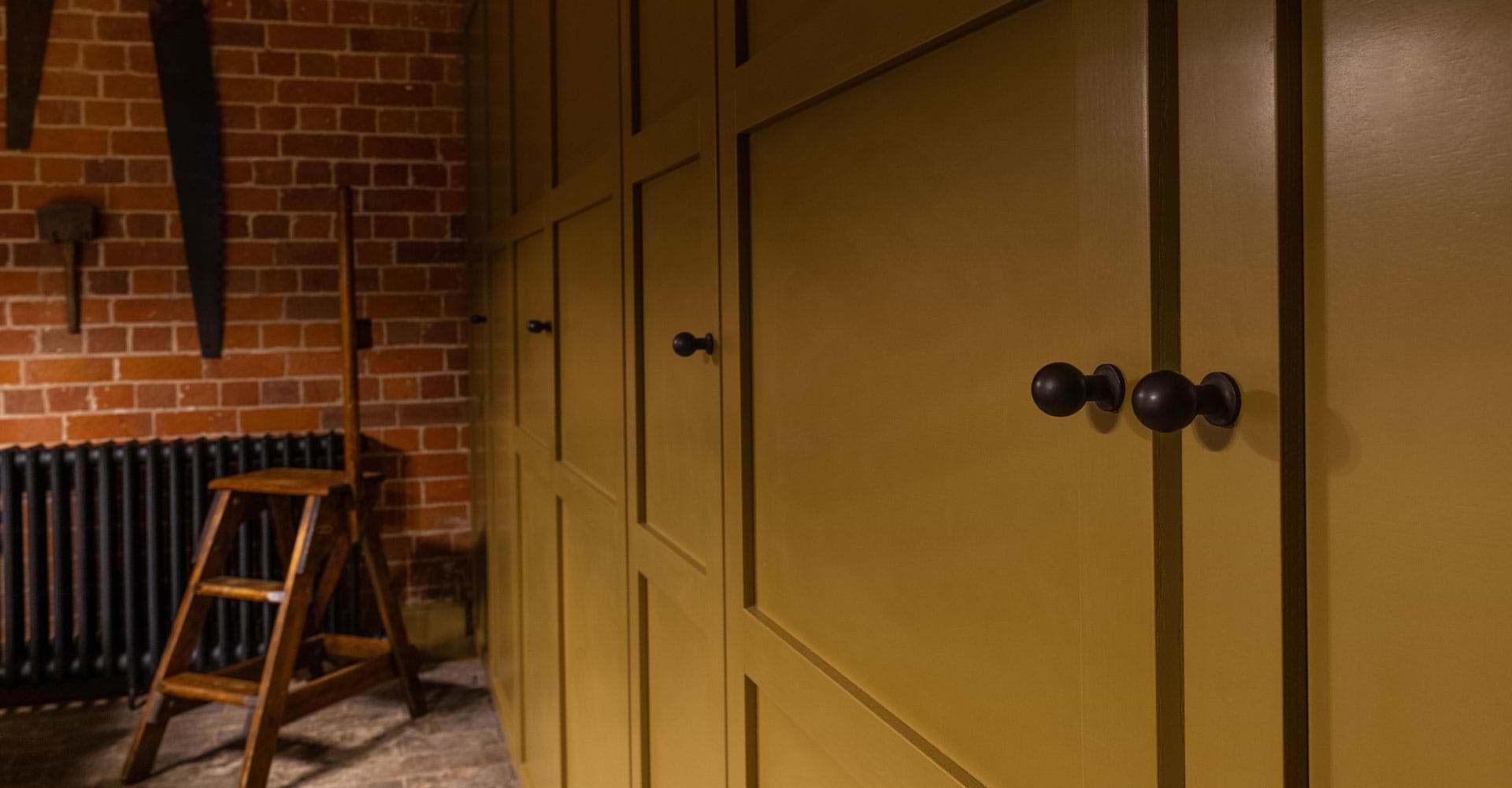 A close-up of olive green cabinets with bronze knobs against a brick wall, featuring a wooden stepladder beside a black radiator.