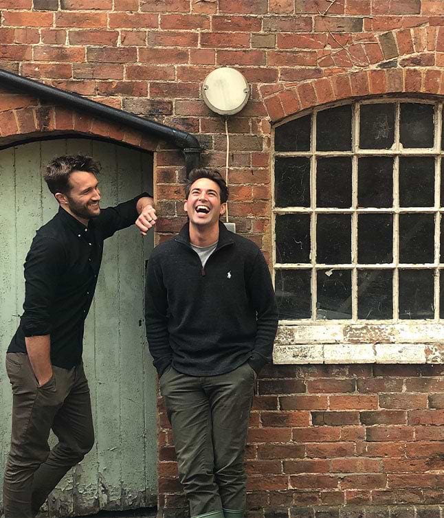 Two young men stand casually against a brick wall, one leaning against a door, with a rustic window visible nearby.