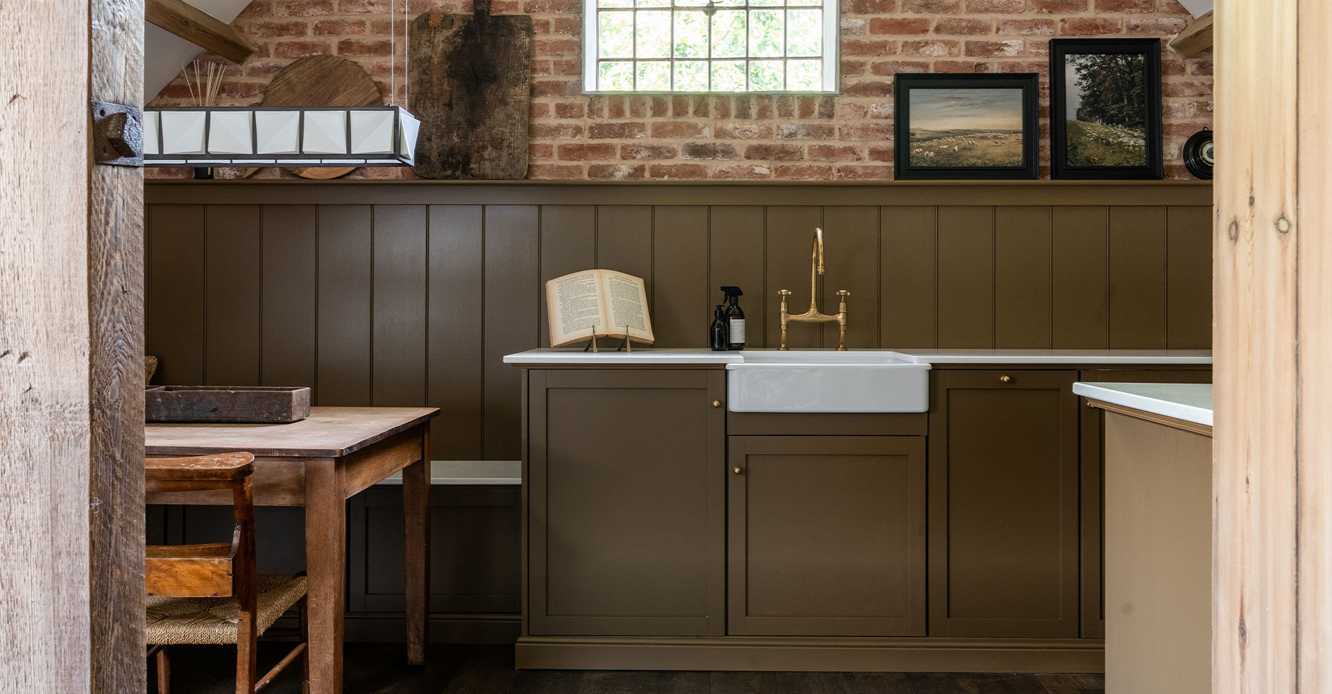A rustic kitchen featuring a brown paneled wall, farmhouse sink, and wooden table, illuminated by a geometric hanging light.