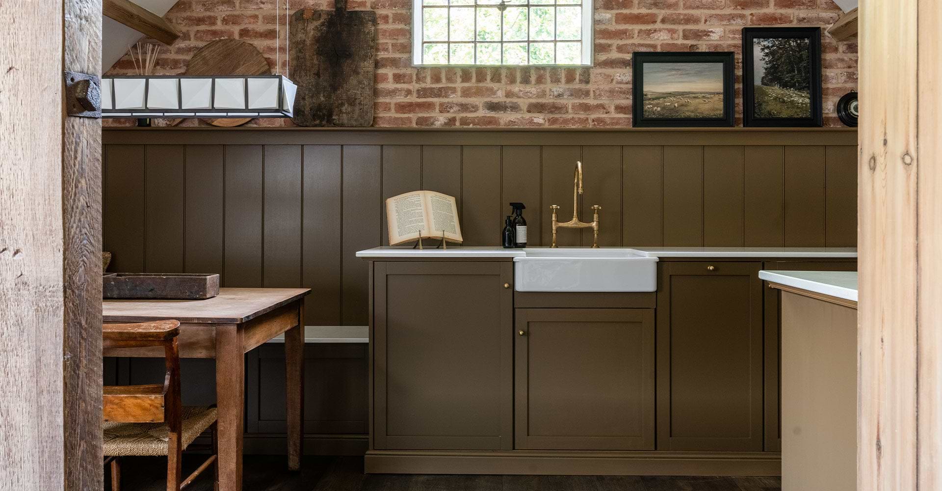 A rustic kitchen featuring a brown paneled wall, farmhouse sink, and wooden table, illuminated by a geometric hanging light.