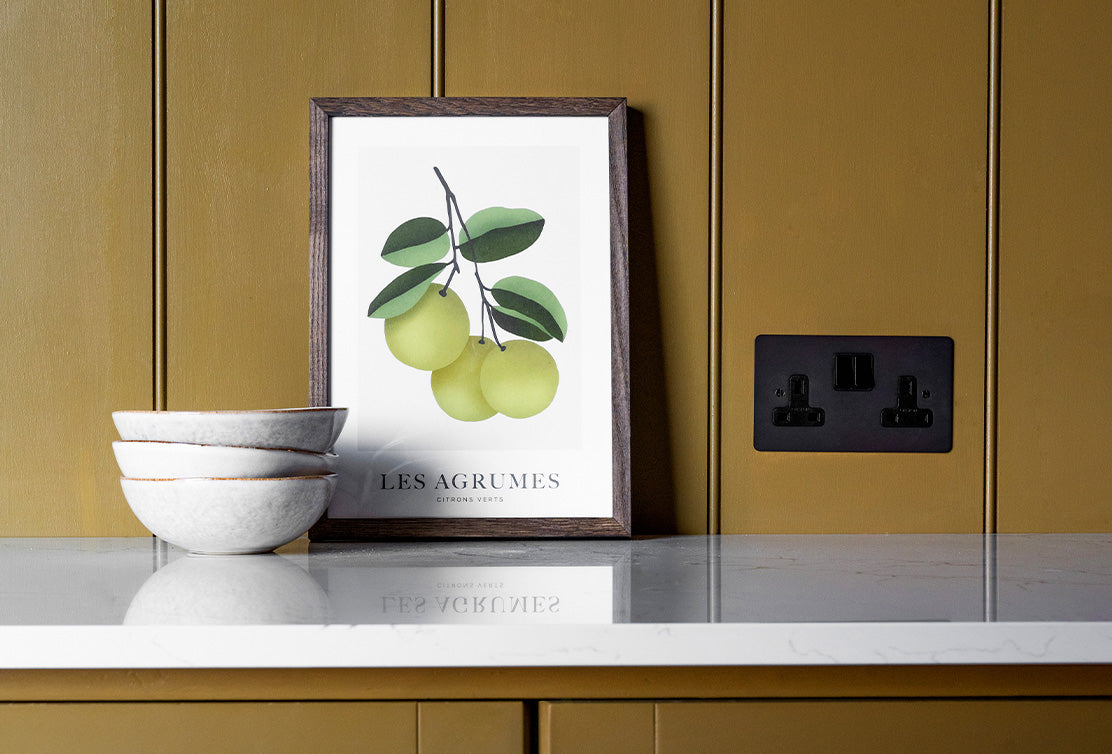 A contemporary kitchen scene featuring a framed print of citrus fruits, three stacked bowls, and a black power outlet against an olive green wall.