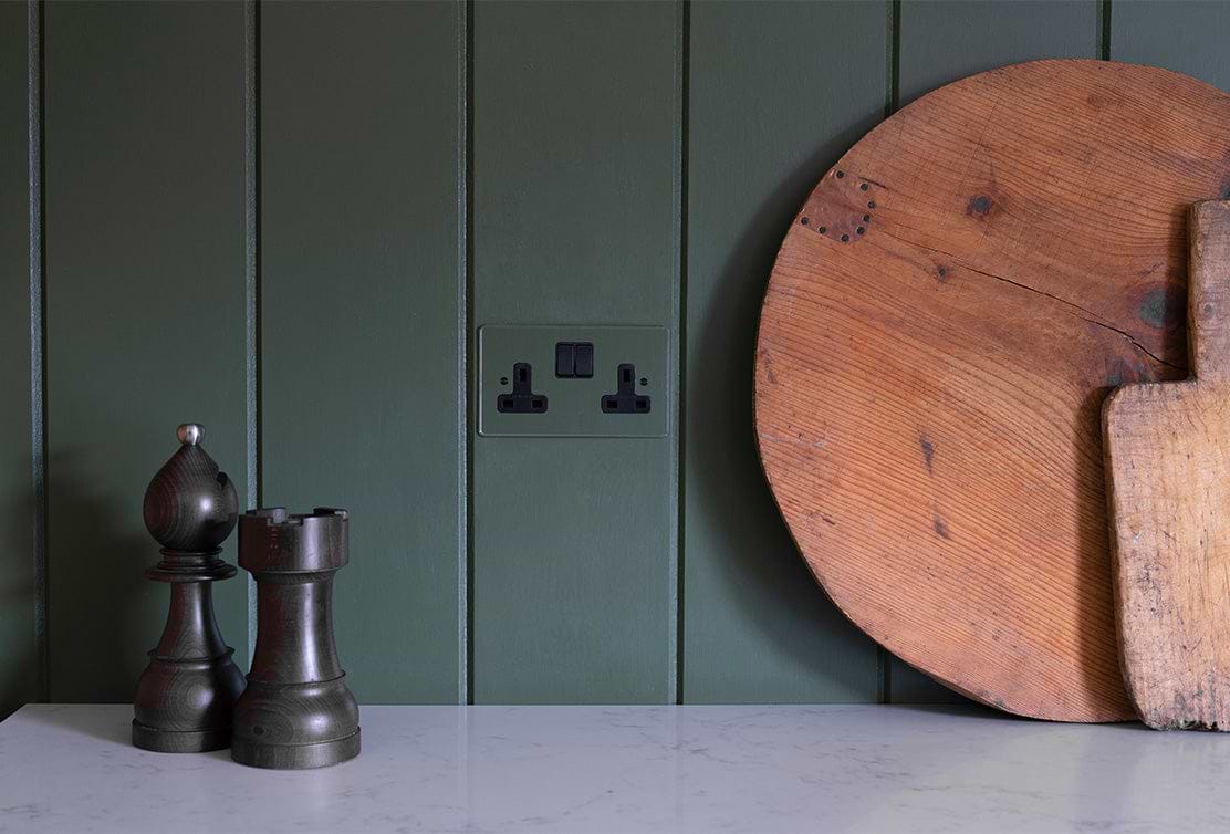 A dark green wall features a power socket, a round wooden cutting board, and two intricately designed wooden chess pieces.