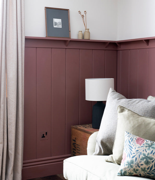 Cozy corner featuring burgundy paneled walls, a white lamp, a wooden crate, and a light sofa with decorative pillows.