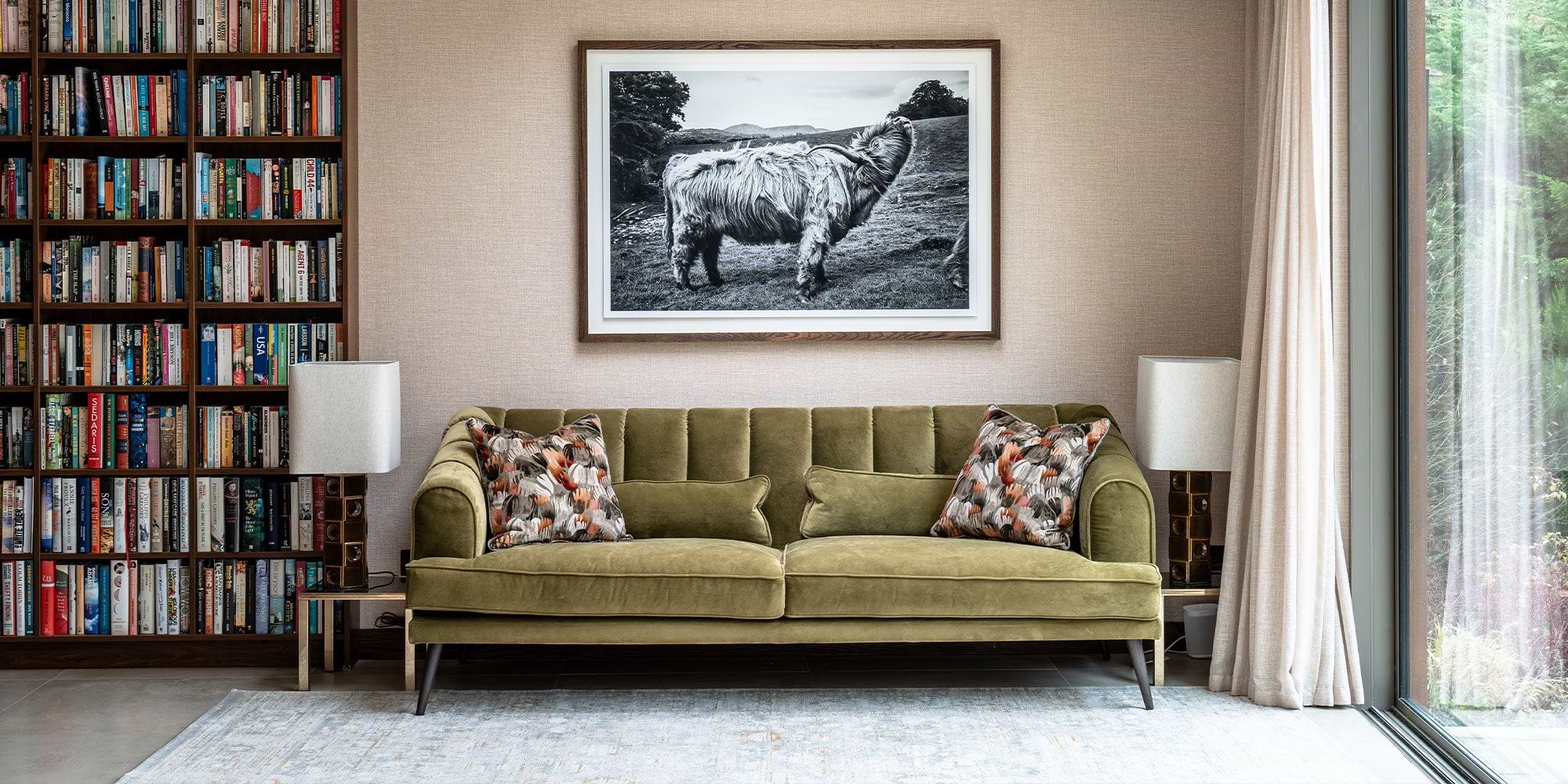 Cozy living room featuring an olive green velvet sofa with decorative pillows, a floor lamp, and a bookcase filled with books. A large black-and-white photo hangs above.