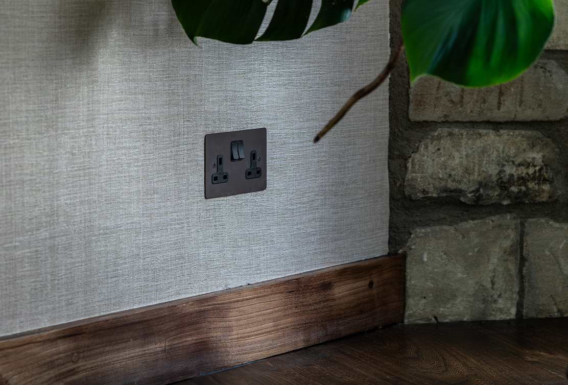 A dark electrical socket plate set against textured beige wallpaper, alongside a wooden baseboard and a green plant leaf overhead.