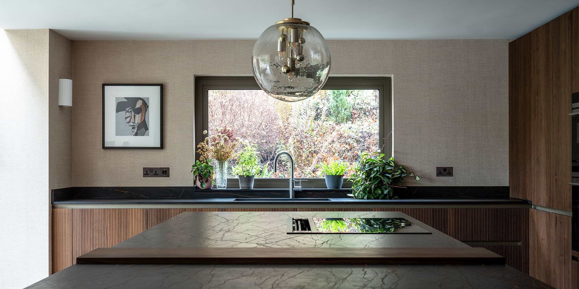 A modern kitchen featuring a large window, a striking pendant light, potted plants, and a stylish marble countertop.