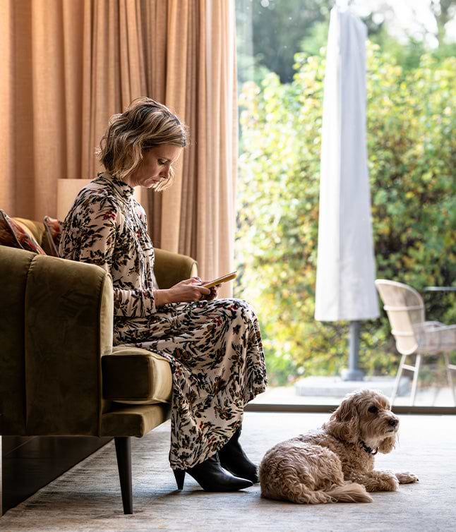 A person in a floral dress sits on a green sofa, holding a phone, while a small dog rests beside her on a cozy rug.