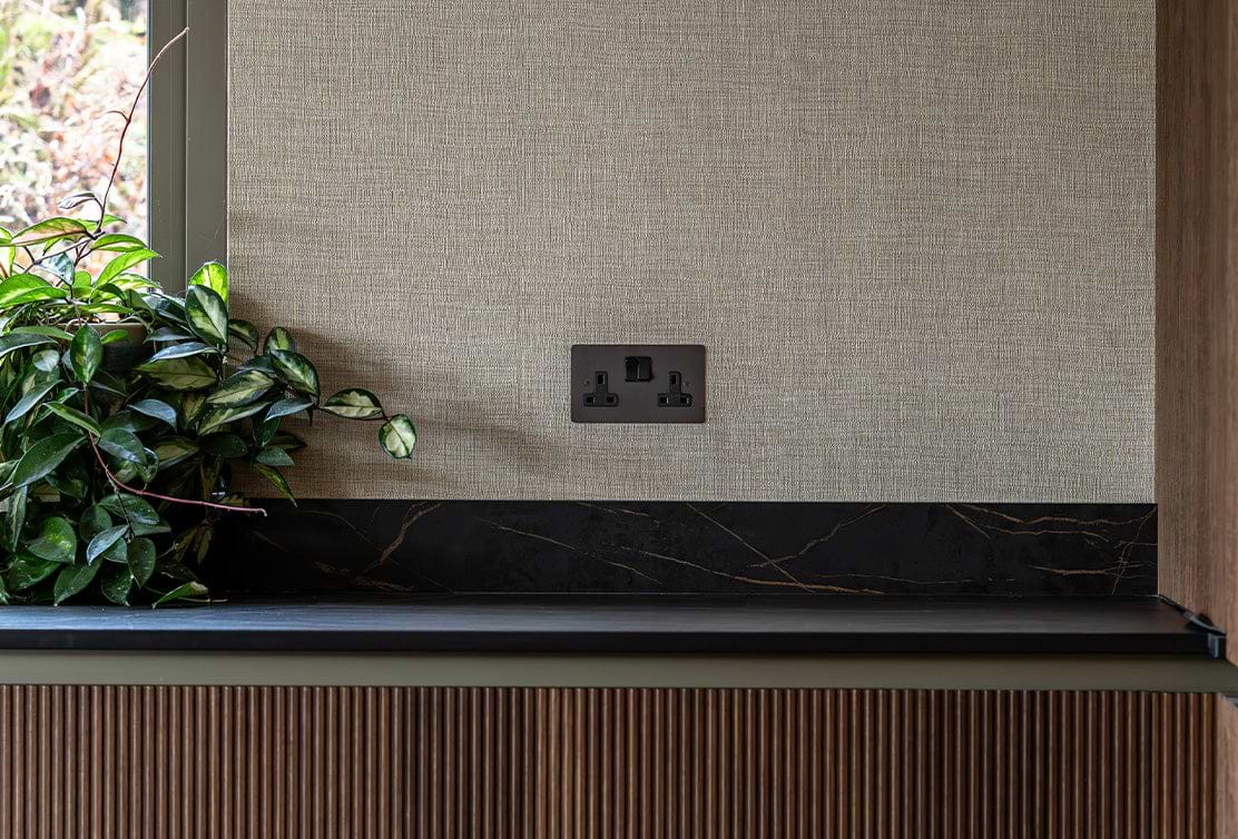 A close-up of a textured wall with a dark electrical outlet, black marble ledge, and green leaves of a plant in the foreground.