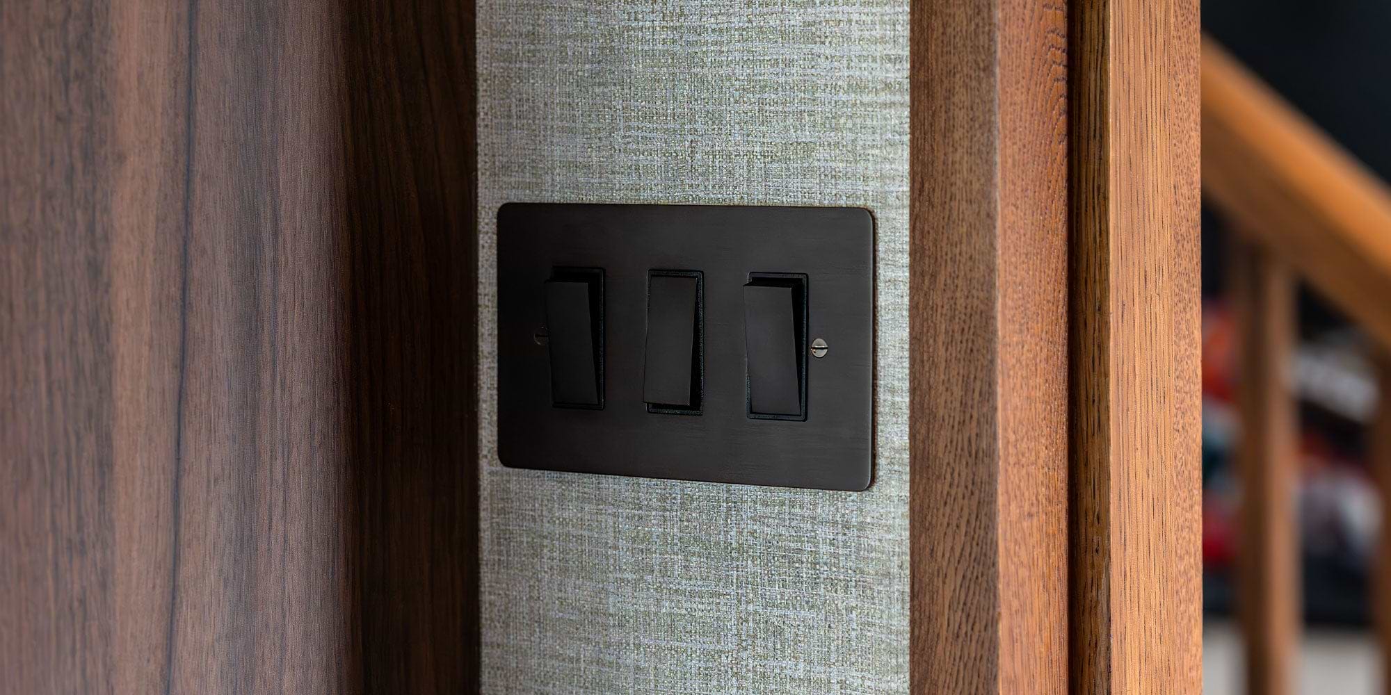 Close-up of a triple light switch with black switches mounted on a textured gray wall, surrounded by wooden trim.