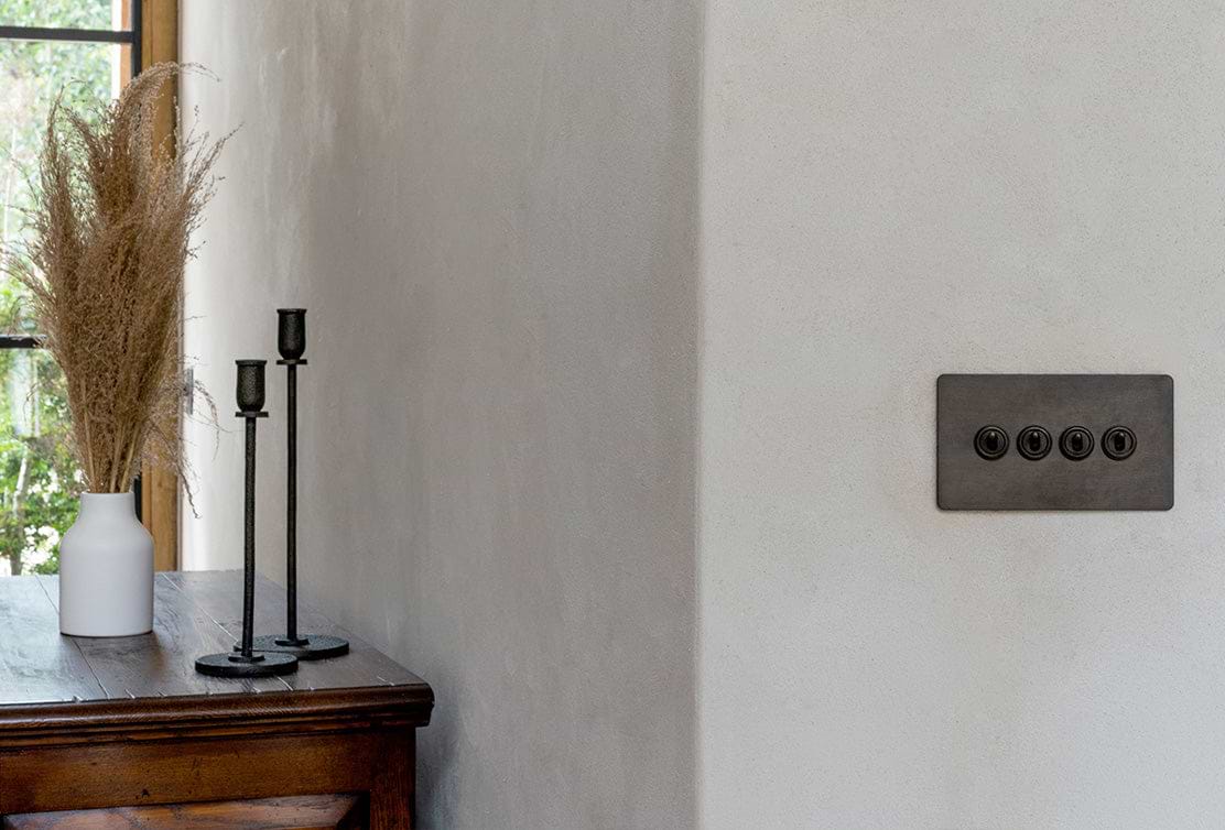 A minimalistic interior featuring a wooden table, black candlesticks, a white vase with dried grass, and a modern light switch on a textured wall.