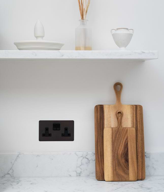 A wooden cutting board leans against a white marble countertop, featuring a bronze electrical outlet and minimalist ceramic items above.