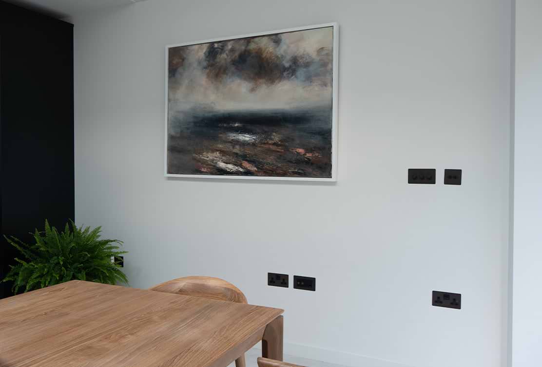 A modern dining area featuring a wooden table, a dark wall, a large abstract painting, and black electrical outlets. A potted plant adds greenery.