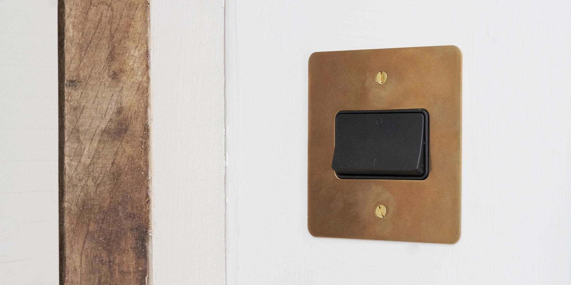 A close-up of a black light switch on a brass plate, mounted on a white wall, with a textured wooden surface visible nearby.