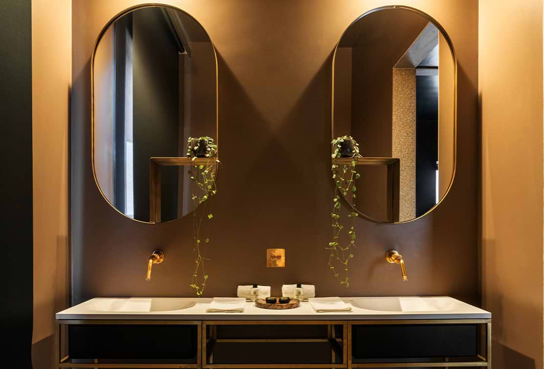 Stylish bathroom featuring dual oval mirrors with gold frames, planters, and a sleek white vanity against a warm brown wall.