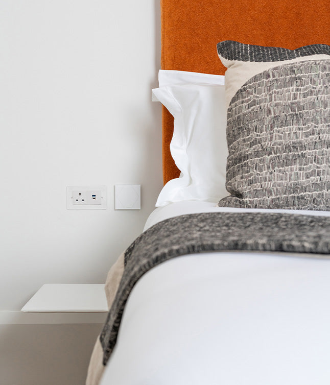 Close-up of a stylish bed with a textured pillow, orange headboard, and side table featuring an electrical outlet.