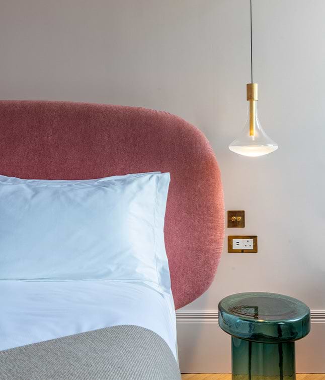 Modern bedroom featuring a pink upholstered headboard, white bedding, a green side table, and a stylish pendant light.