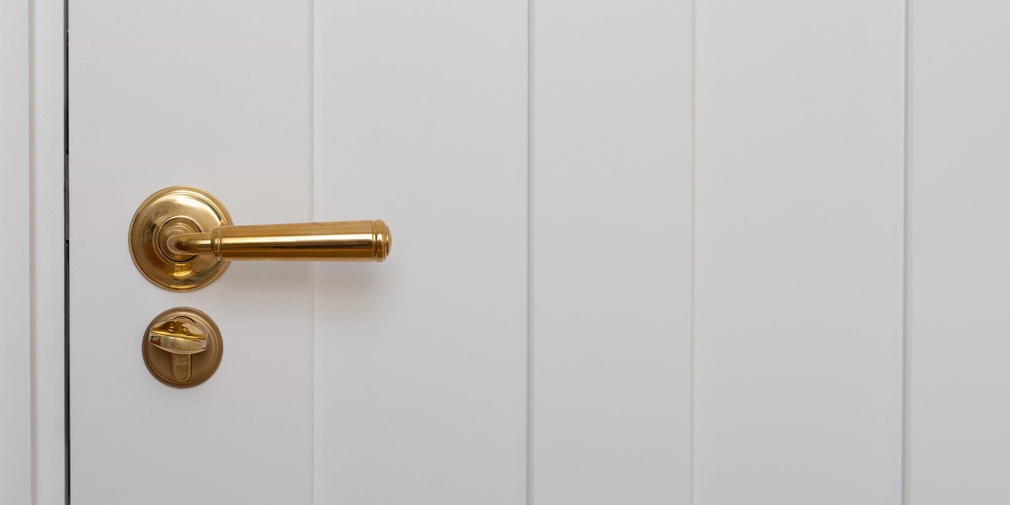 A close-up of an antique brass door handle and thumb turn on a white paneled door. The handle is sleek and shiny against the plain background.