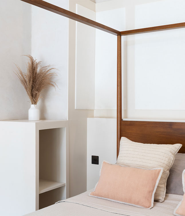 A cozy, minimalist bedroom featuring a wooden bed frame, soft pillows, and a decorative vase with dried grass against a light wall.