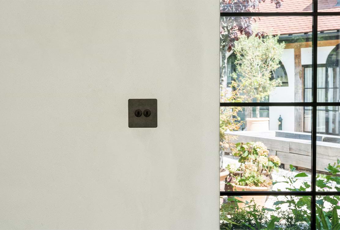 A close-up of a modern black light switch on a smooth white wall, with a garden visible through a large window beside it.