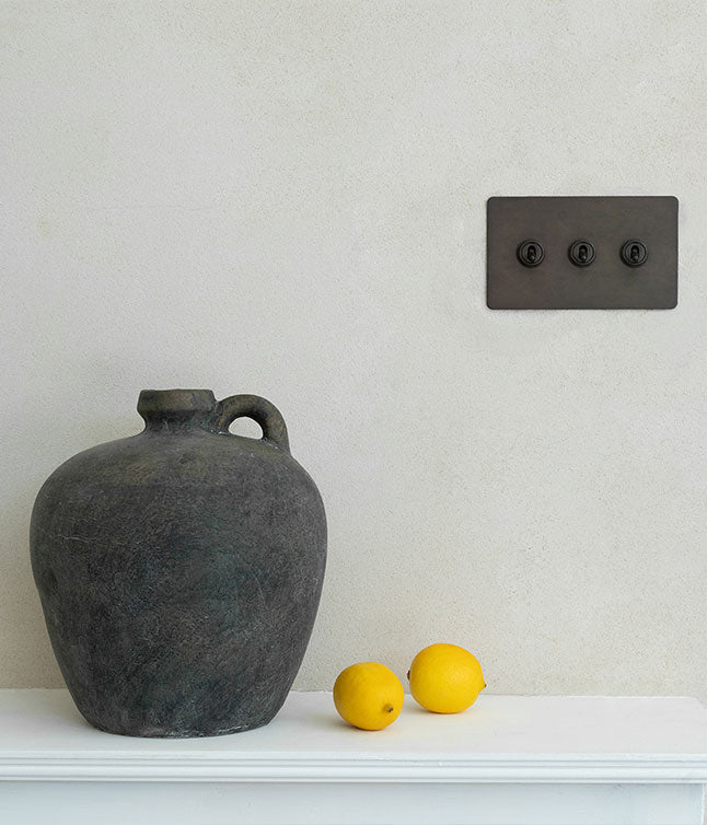 A textured dark ceramic vase beside two fresh yellow lemons on a white shelf, against a smooth light-colored wall.
