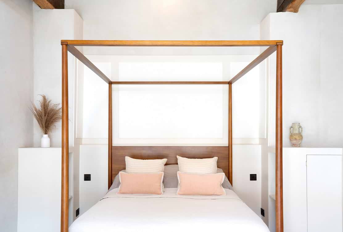 A cozy, modern four-poster bed with neutral pillows against a light, minimalist backdrop, accented by decorative plants nearby.