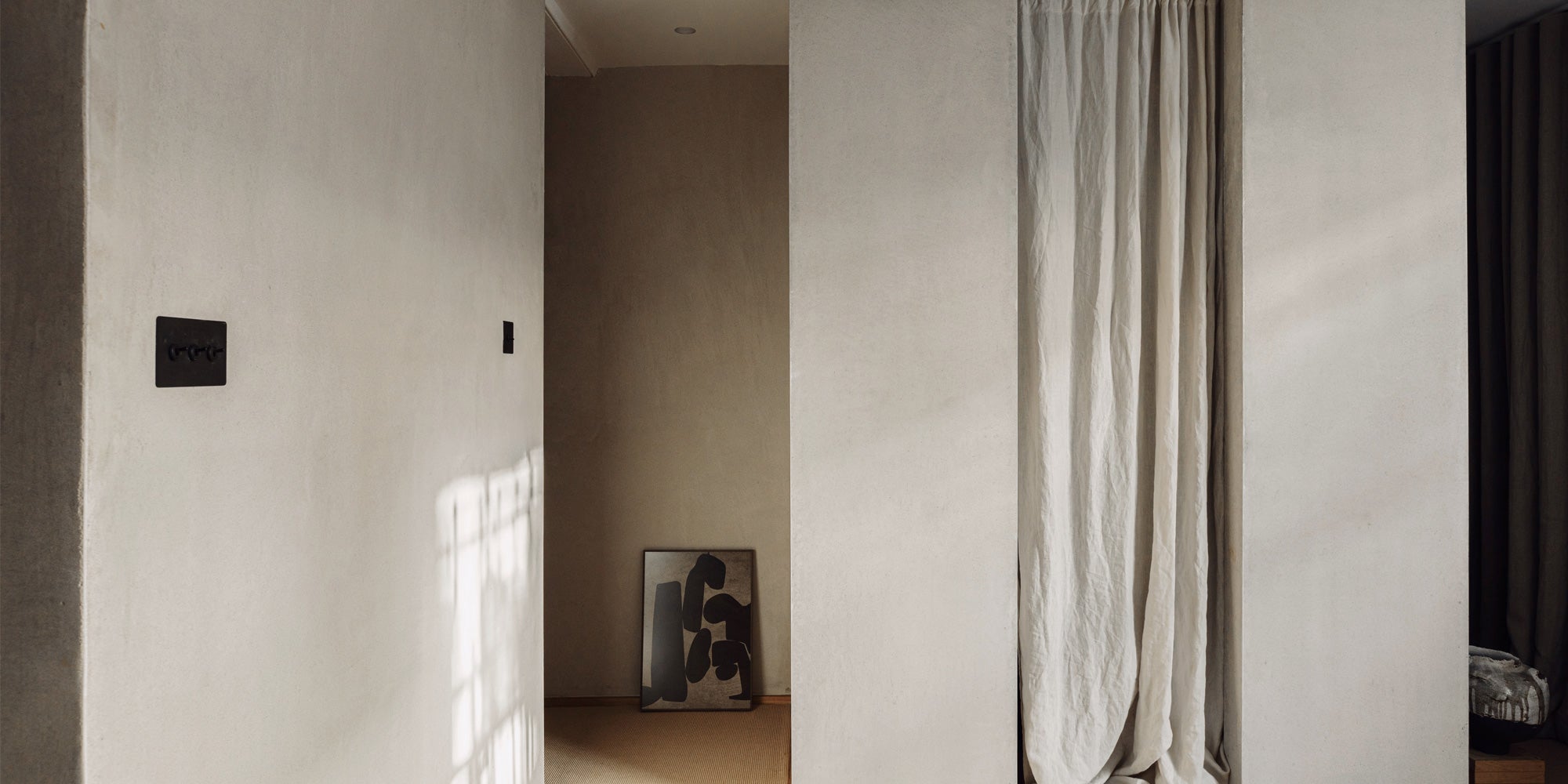 A neutral-toned hallway features a textured wall, a black light switch, and an abstract black artwork against a soft beige background.