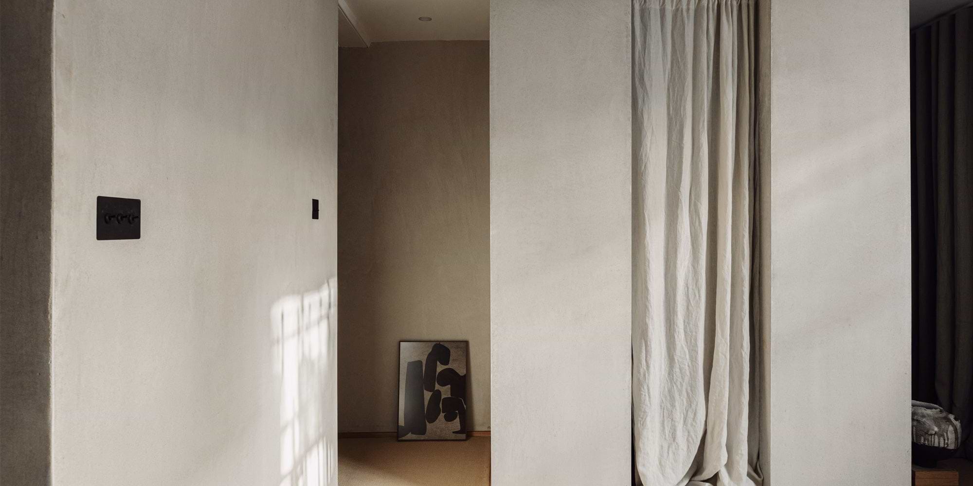 A neutral-toned hallway features a textured wall, a black light switch, and an abstract black artwork against a soft beige background.