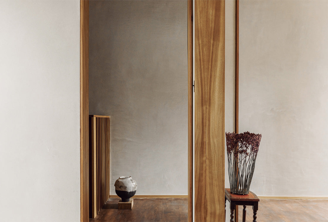 A minimalist interior featuring a wooden column, a rustic pot on a pedestal, and a vase of dried flowers on a small table.