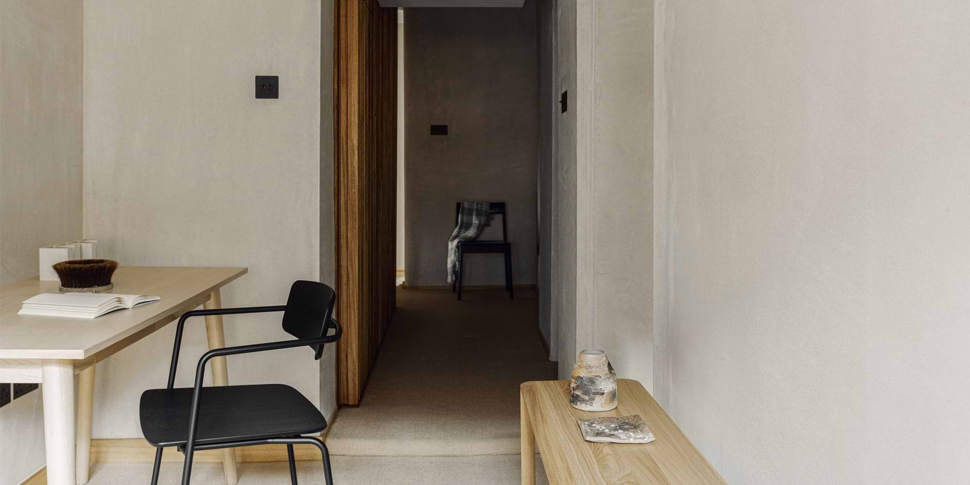 A minimalist hallway featuring a wooden table, a black chair, and a chair with a plaid throw, all against soft, neutral-toned walls.