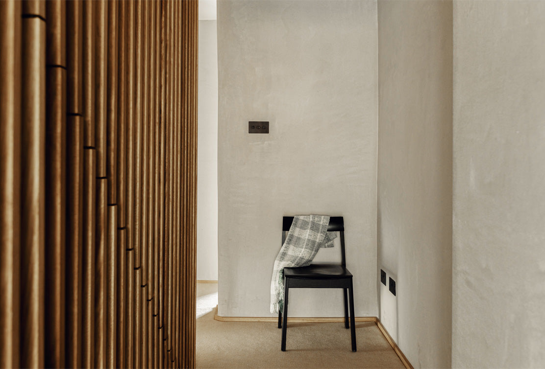 A minimalist hallway featuring a wooden slat partition, a black chair with a gray checkered blanket, and a neutral-colored wall.