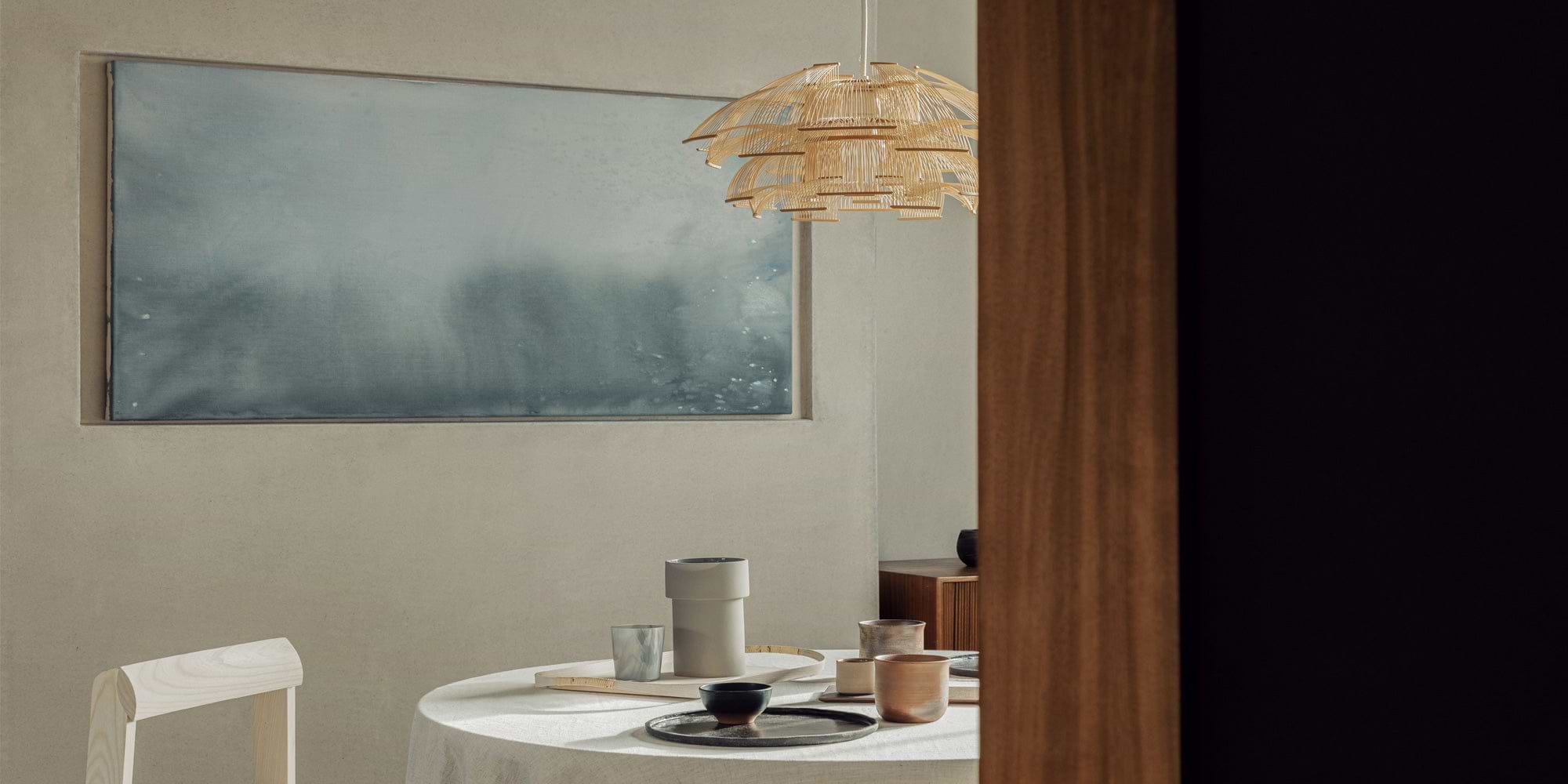 A modern dining space with a round table set with minimalist plates and cups, illuminated by a stylish bamboo pendant light.