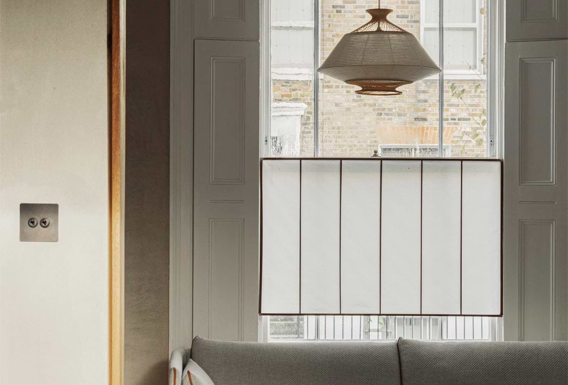 A cozy living space featuring a gray sofa, a woven pendant light, and a white window shade, with sunlight filtering through.